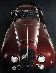 the front end of an old red car with its hood up and lights on, against a black background