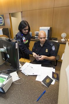 two police officers sitting at a desk looking at papers on the computer screen while another officer stands in front of them