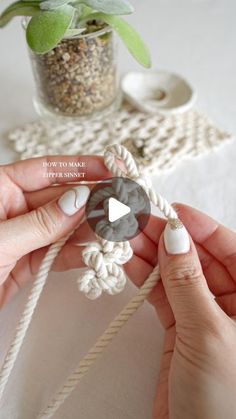 two hands are holding a piece of white yarn with black and gray buttons on it, next to a potted plant