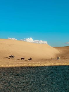 three people riding horses on the sand dunes near a body of water in front of a blue sky