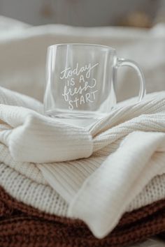 a glass coffee mug with the words today is a fresh start sitting on top of a pile of sweaters