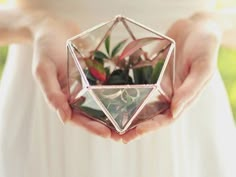 a person holding a glass vase with plants in it's hands, while wearing a white dress