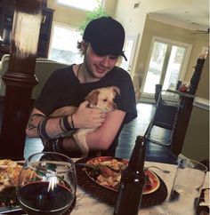 a man holding a small dog in his arms while sitting at a table with food and wine