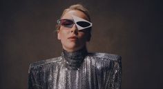 a woman with an eye patch on her face wearing a silver jacket and sunglasses, standing in front of a dark background