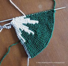 a green and white knitted triangle hanging from a hook on a wooden table with knitting needles