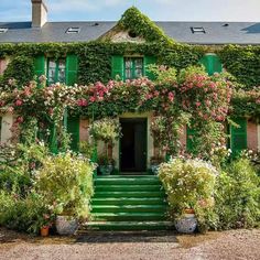 an old house with green shutters and flowers on the front door, surrounded by greenery