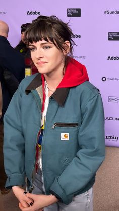 a woman standing in front of a purple wall wearing a blue jacket and red hoodie