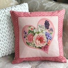 a pink heart pillow with flowers on it sitting on a couch next to two pillows