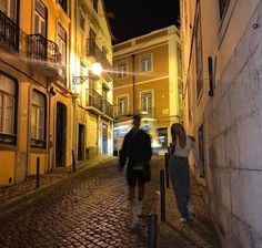 two people are walking down an alley way at night in front of some old buildings