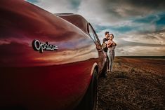 a man and woman standing next to a red car in the middle of a field