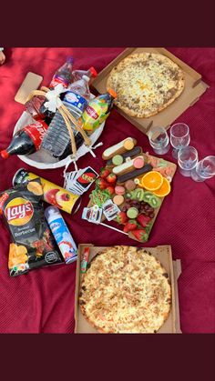 two pizzas, drinks and snacks on a bed with red sheets in the background