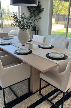 a dining room table with white chairs and plates on the placemats, in front of a large window