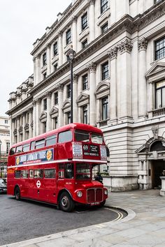 a red double decker bus driving down a street next to a tall building with many windows