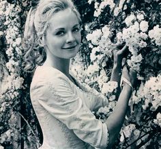 a woman in a white dress standing next to some flowers and holding onto a plant