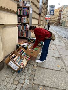 a woman leaning over a box filled with books on the side of a street next to a book shelf