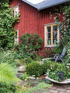 a red house surrounded by greenery and flowers with a bench in the foreground