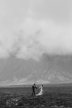 a bride and groom holding hands in front of a mountain range with clouds rolling over them