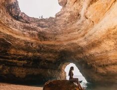 a woman sitting on top of a large rock near the ocean in front of a cave