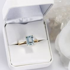 an engagement ring with a blue topaz sits in a white box next to flowers