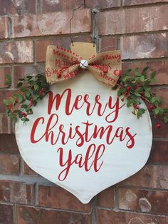 a merry christmas y'all sign hanging on the side of a brick wall with holly and burlap