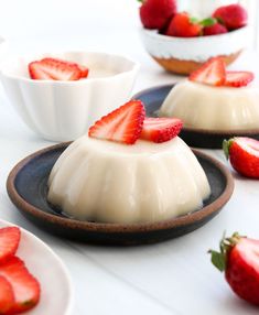 three desserts with strawberries sit on plates next to bowls and spoons in the background