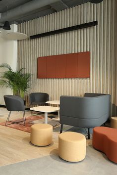 An interior of Nowy Styl showroom. Meeting zone furnished with grey Tepee sofa and two grey Naudu armchairs and couple of yellow and red Tapa pouffes. Wooden and concrete floor. Wooden lamels on the wall and several red acoustic wall panels Sileo. Palma Areca in the background. Modern Office Lounge, Small Show Room Ideas, Cosy Office Design, Paint Zoning, Office Hallway Decor, Colorful Conference Room, Office Chill Area, Warm Office Design, Wellness Office