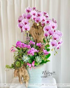 a vase filled with lots of purple flowers on top of a white table next to a curtain