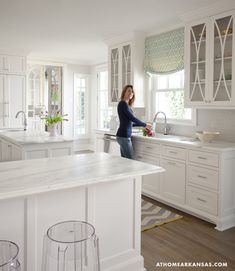 a woman standing in the middle of a kitchen with white cabinets and marble counter tops