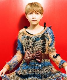 a young boy in a blue and brown dress standing against a red wall with his hands on his hips