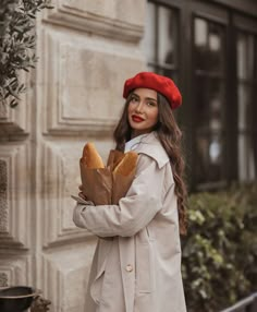 a woman wearing a red hat and trench coat holding bread in front of a building