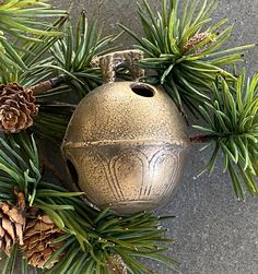 a metal bell surrounded by pine cones and evergreen needles on a concrete surface in the daytime