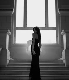 a woman in a long black dress standing on some stairs with her hands behind her back