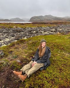 a woman sitting on top of a lush green field next to a mountain covered in clouds