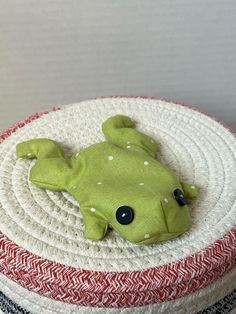 a green stuffed frog sitting on top of a white and red cushion with black eyes