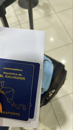 a person holding up a blue and yellow passport in their left hand while standing next to luggage