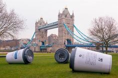 two trash cans sitting on the grass in front of a tower bridge with trees and buildings in the background