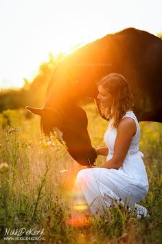 a woman kneeling down next to a black horse in a field at sundown with the light shining on her face