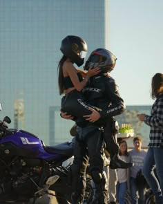 two people on a motorcycle are hugging each other while others stand around and watch them