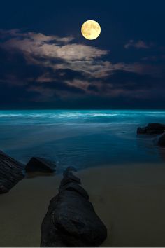 a full moon is seen over the ocean with rocks in the foreground and dark clouds