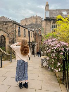 a woman standing on the sidewalk in front of some buildings and looking up at flowers