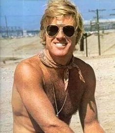 a shirtless man sitting on the beach with his hands in his pockets and wearing sunglasses