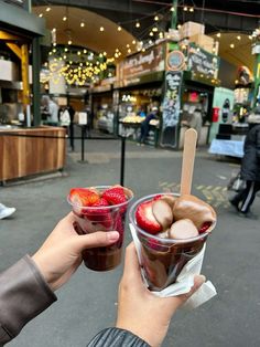 two people holding up cups with ice cream and strawberries in them on the street