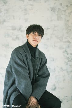 a young man sitting on top of a wooden stool wearing a coat and black pants