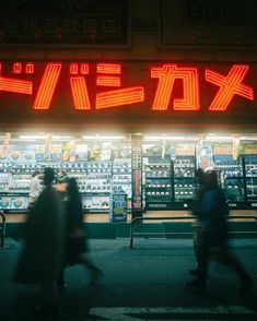 people walking past a convenience store with neon signs on the front and side windows at night