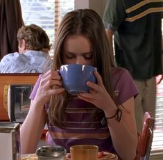 a woman sitting at a table drinking from a blue cup and holding a coffee mug in front of her face