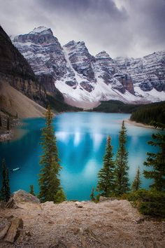 a lake surrounded by snow covered mountains and pine trees in the foreground, under a cloudy sky