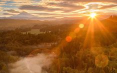 the sun shines brightly over a forest with mountains in the background and fog rising from the ground