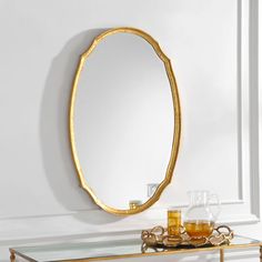 an oval mirror sitting on top of a table next to a glass vase and two glasses