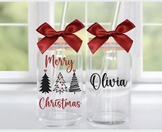 two clear glass bottles with red bows and merry christmas lettering on the side, sitting next to each other in front of a window