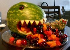 a watermelon carved to look like a shark with its mouth open and fruits in the foreground